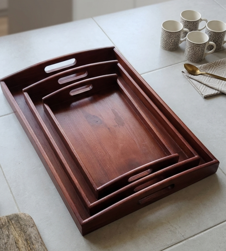 Three nested rectangular wooden serving trays with cutout handles, displayed on a light stone countertop with ceramic mugs and a spoon nearby.