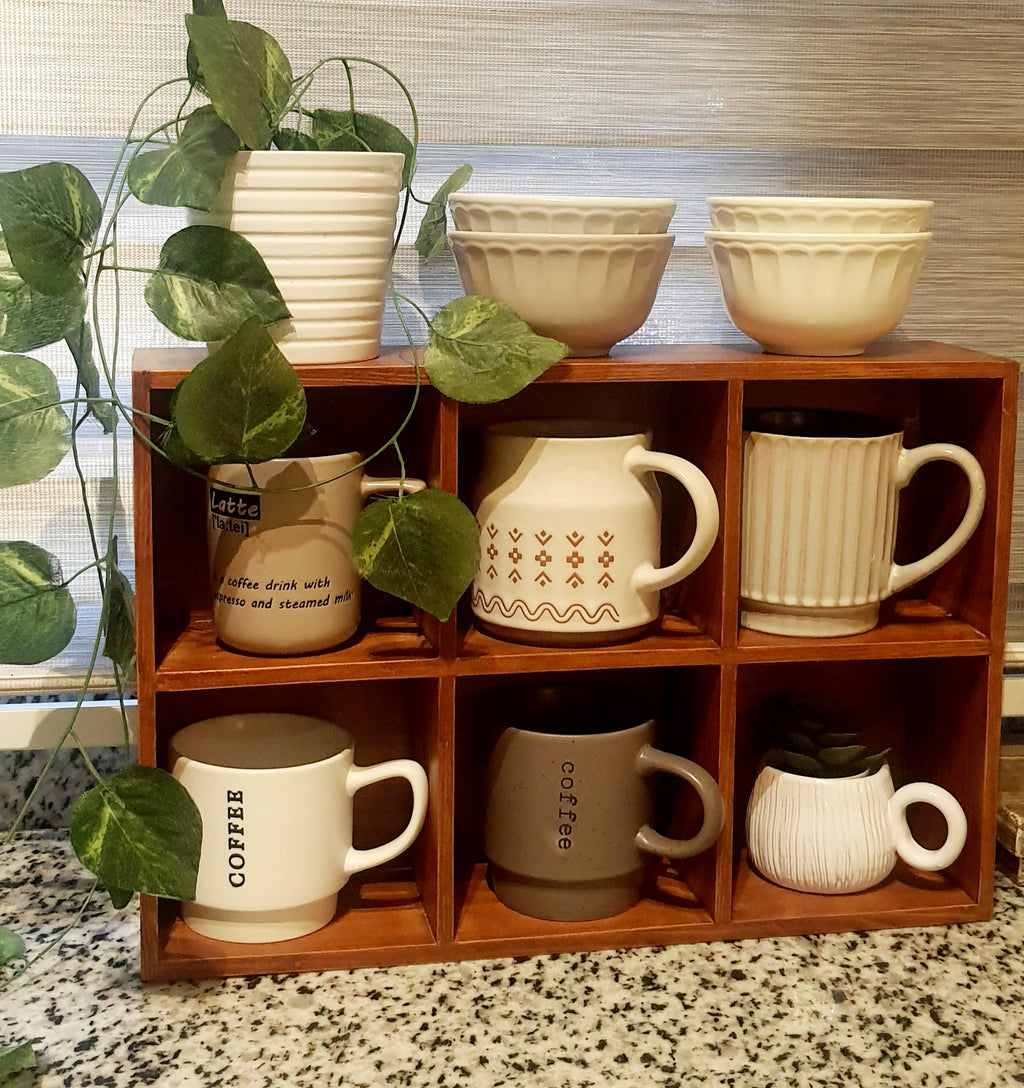 Collection of ceramic mugs and bowls on a wooden shelf with plants around.