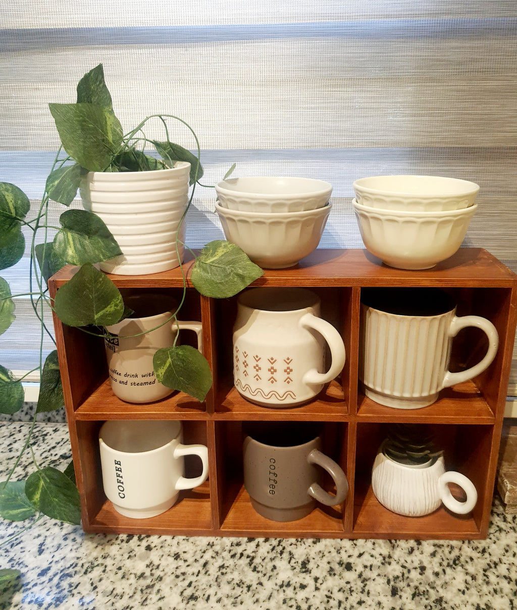 Set of ceramic mugs and bowls on a wooden shelf with a plant in the background.