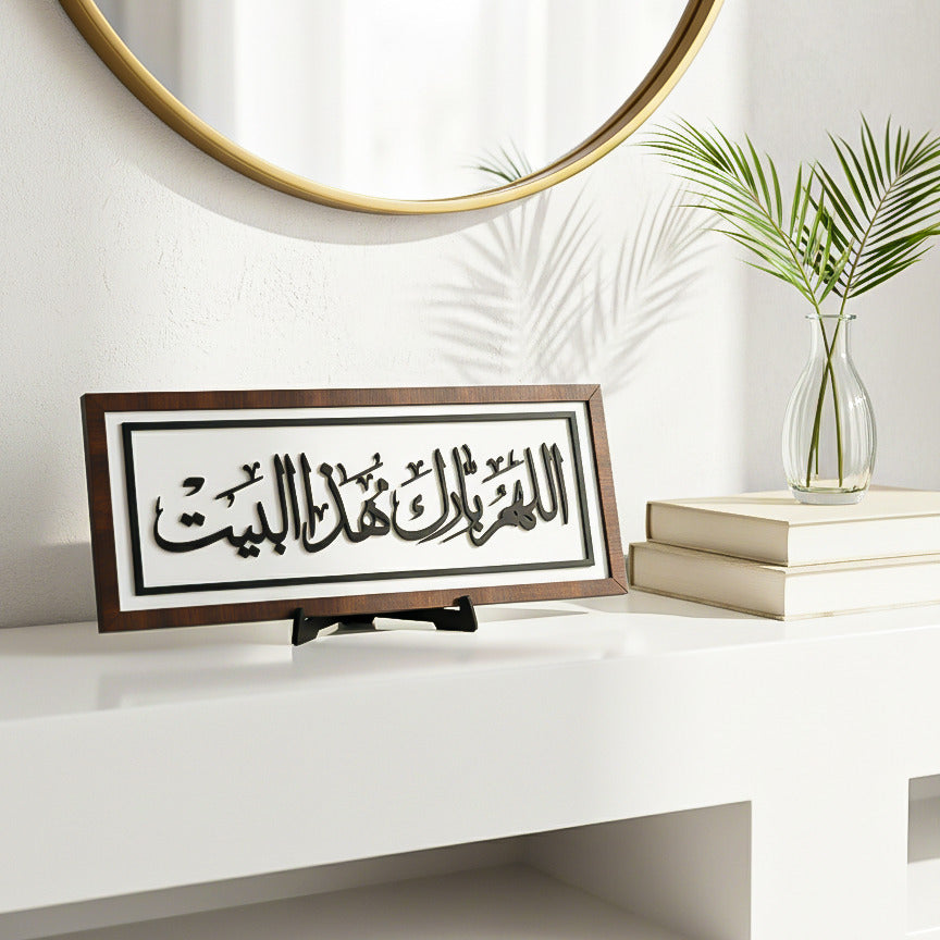 Decorative frame with Arabic calligraphy on a white surface with a plant and books in the background.