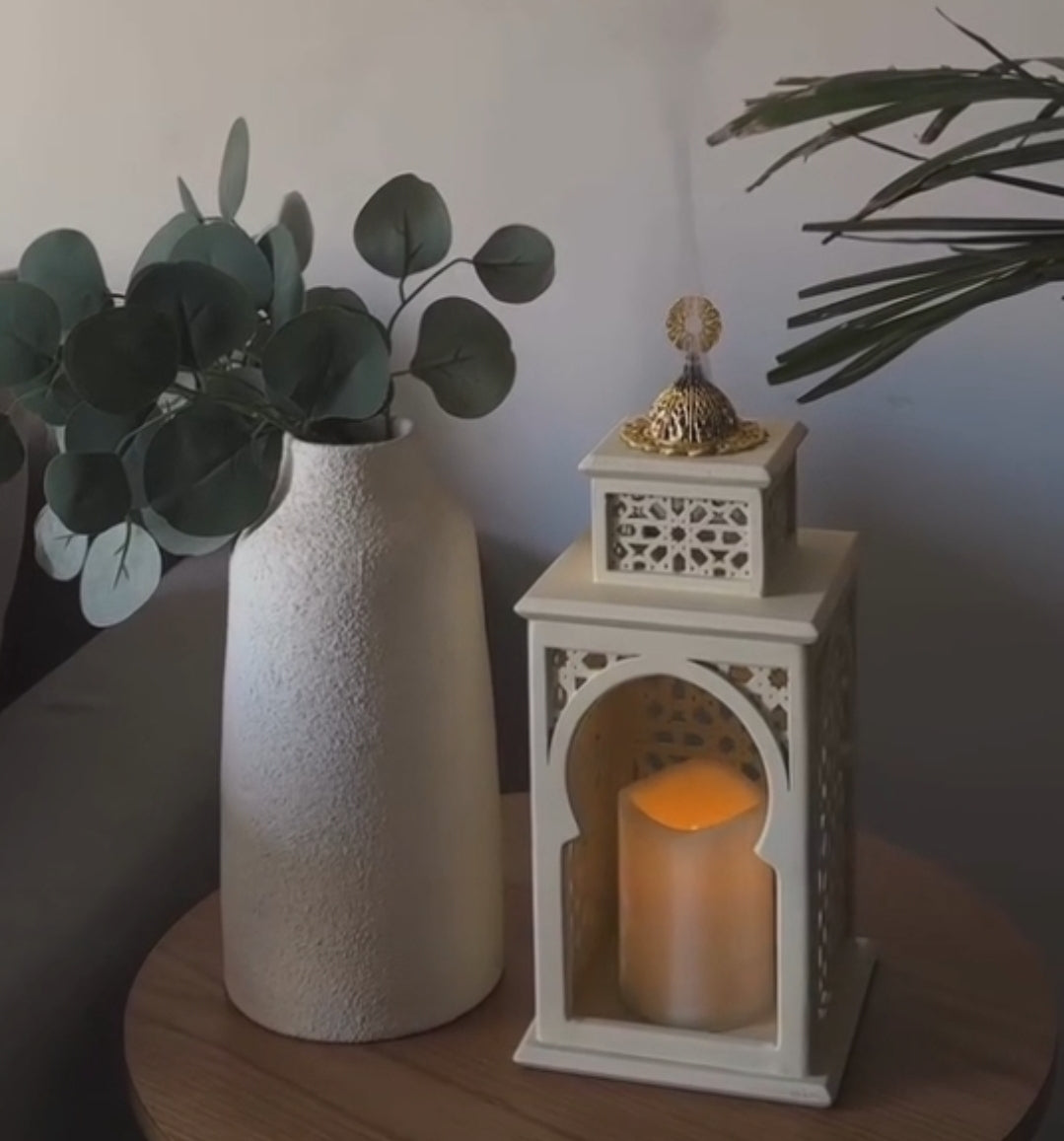 Decorative scene with a textured vase, eucalyptus leaves, and a white lantern with a candle on a wooden surface.