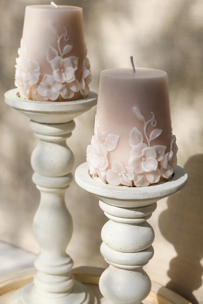 Two pink candles with floral designs on white candlesticks against a blurred natural background.